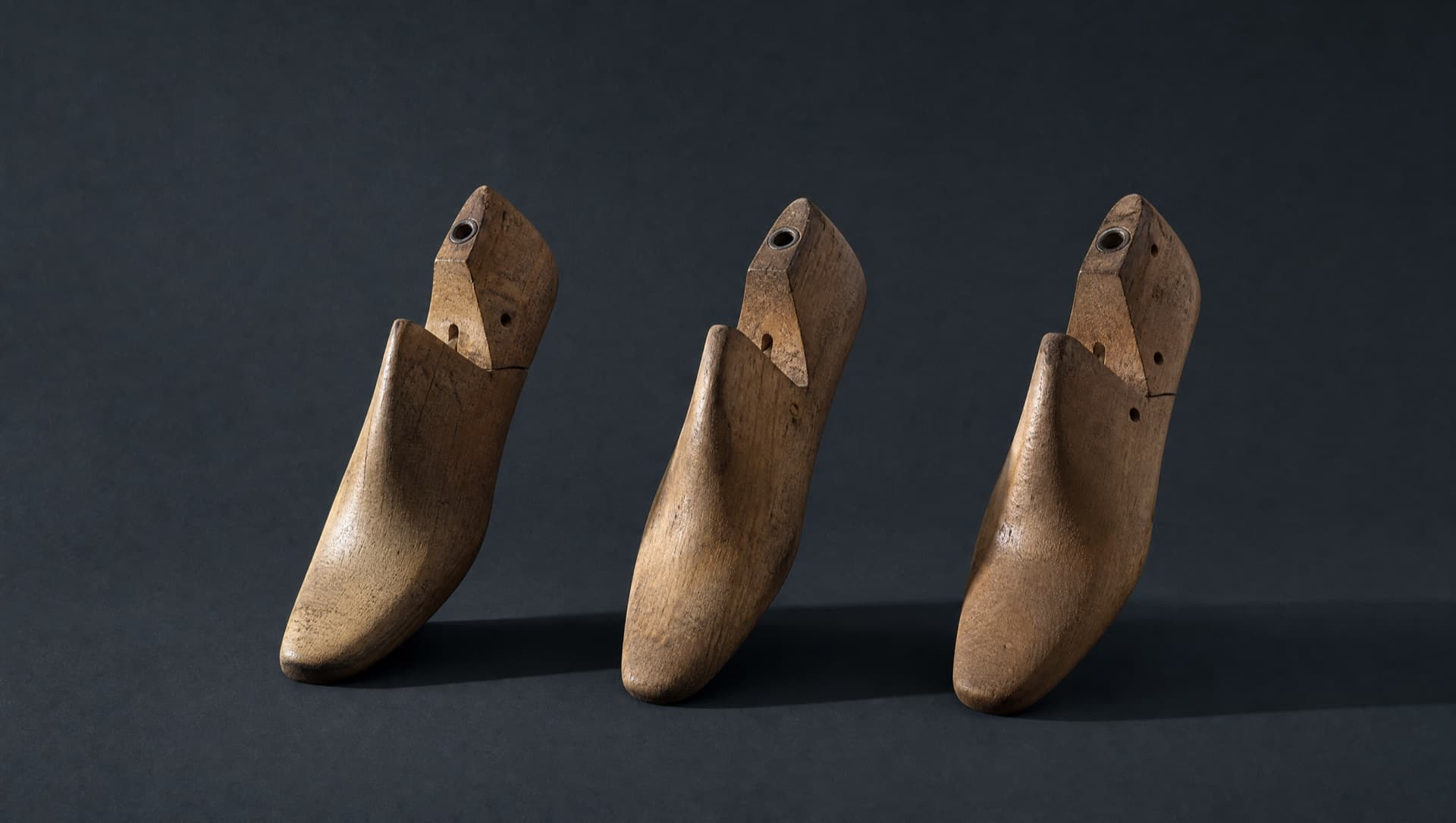 Three wooden shoe lasts standing in a row on a dark slate ground, lit by one hard raking light casting long parallel shadows.