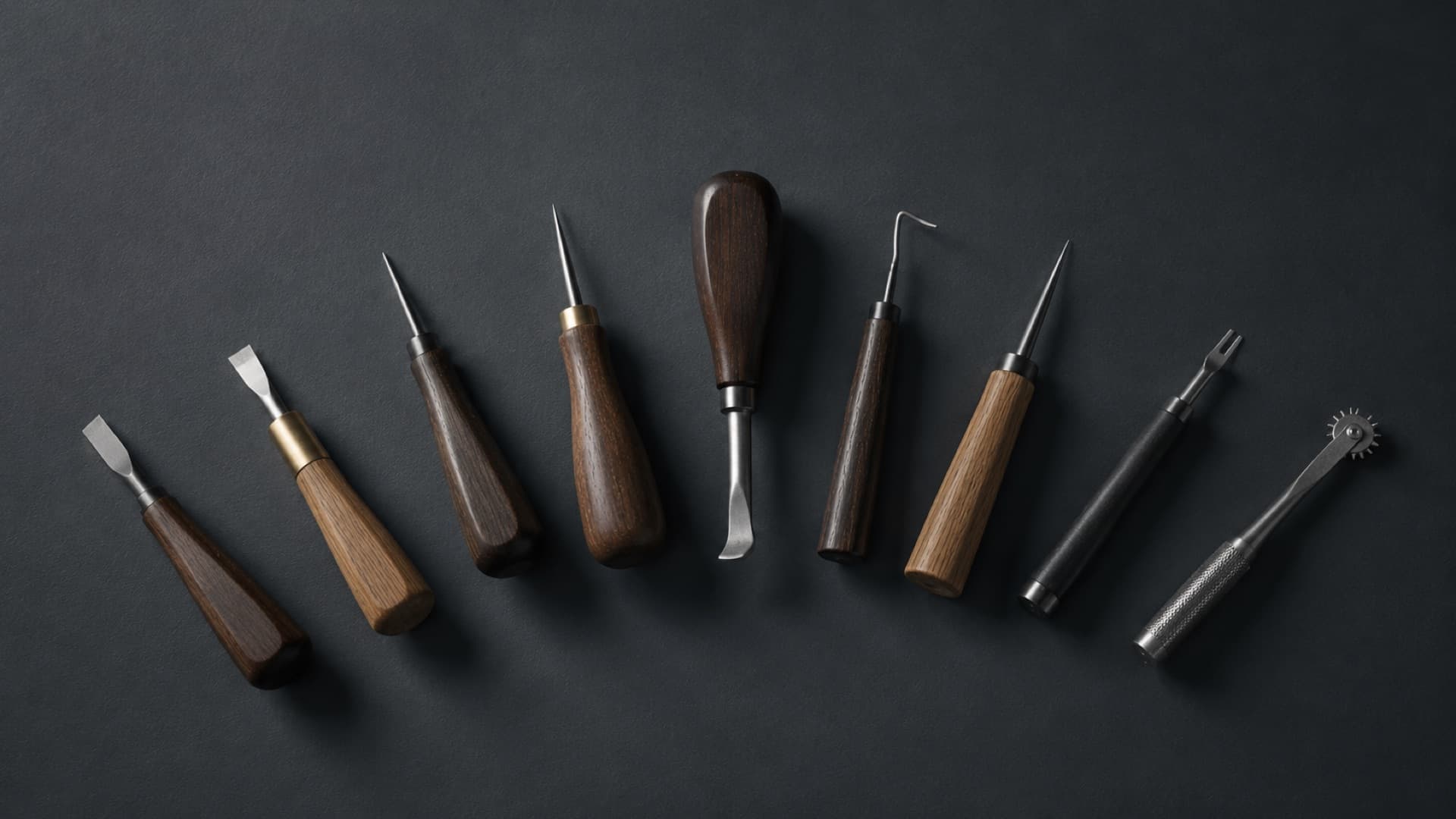 Nine small shoemaking hand tools laid out flat in a shallow arc on a dark slate ground.